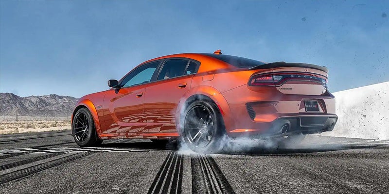 Orange Dodge Charger with wide body kit doing a burnout, smoke rising from its tires on an asphalt track in a desert landscape.