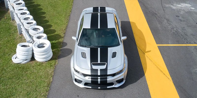 Aerial view of a silver muscle car with black racing stripes on a racetrack, next to a tire barrier and yellow lines.