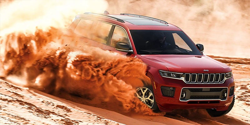 Red Jeep Grand Cherokee SUV driving fast through sand, kicking up a large cloud of dust.