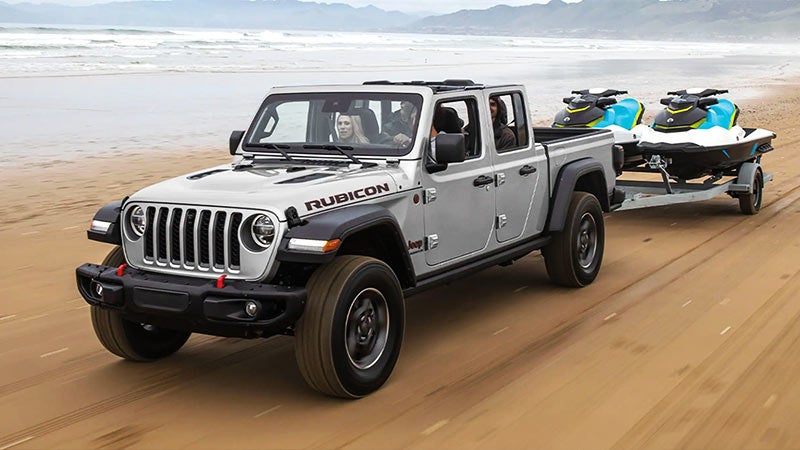 Silver Jeep Gladiator Rubicon pickup truck on a sandy beach, towing a trailer with two blue and black jet skis.