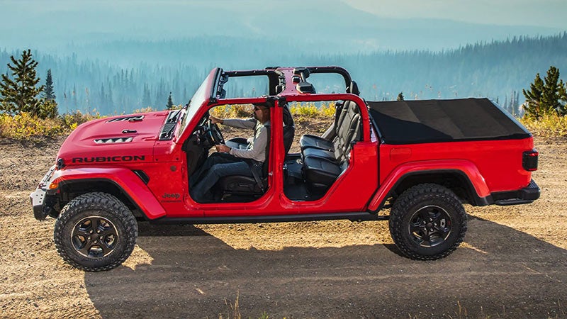 Red Jeep Gladiator Rubicon with doors removed, soft top over bed, a man driving on a dirt road with mountains behind.