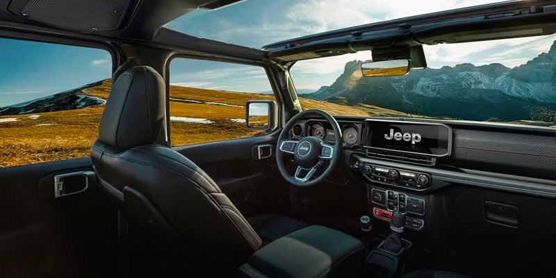 Interior of a Jeep Wrangler, black seats, dashboard, and open top, with mountains and golden hills visible outside.