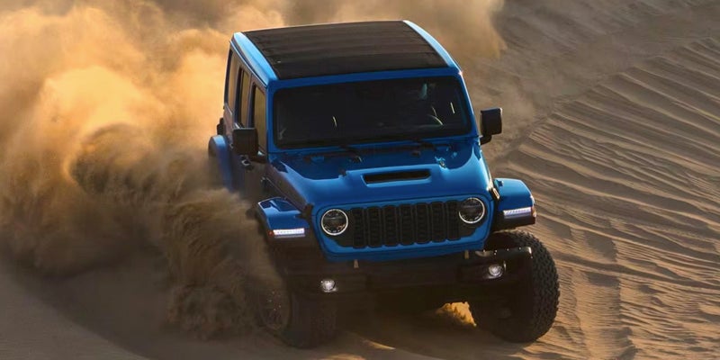 Blue Jeep SUV driving fast through sand dunes, kicking up a large cloud of dust.